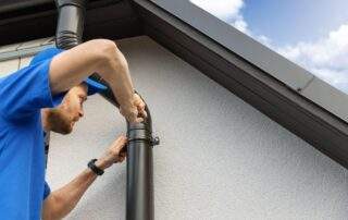 A professional in a blue shirt and baseball cap is installing a new brown gutter downspout on the side of a modern house. He is using a screwdriver to fasten a clamp around the downspout. The house has a white, textured exterior and a dark brown trim. The sky is blue with scattered clouds.