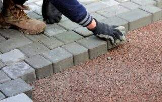 Close-up of a worker in gloves laying a border of gray concrete paver patio stones on a gravel base, showing the construction and future maintenance needs of a paving project.